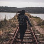 A woman walks on railroad tracks next to a serene lake under a cloudy sky, capturing a sense of adventure and solitude.